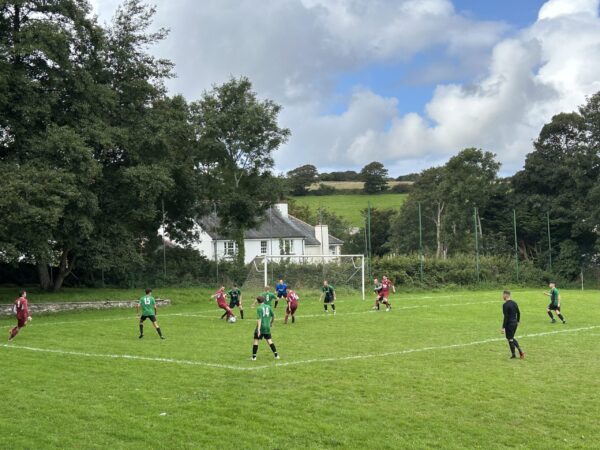 Village football match on Chacewater Recreation Ground pitch; maroon side attacks while green team defends near goal amid trees and cottages.