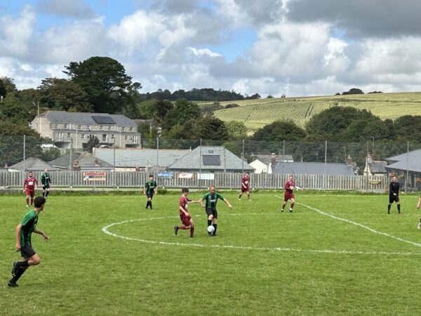 Local amateur football match at Chacewater Recreation Ground, players in maroon and green kits under cloudy sky, countryside hills in background