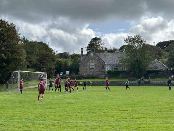 Amateur football match: maroon team defends corner near goal on Chacewater Recreaton Ground pitch, Chacewater School in background under cloudy sky.