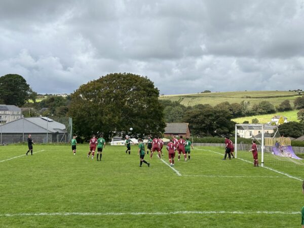 Amateur football corner kick on Chacewater Recreation Ground pitch: maroon vs green near goal, cloudy countryside backdrop.